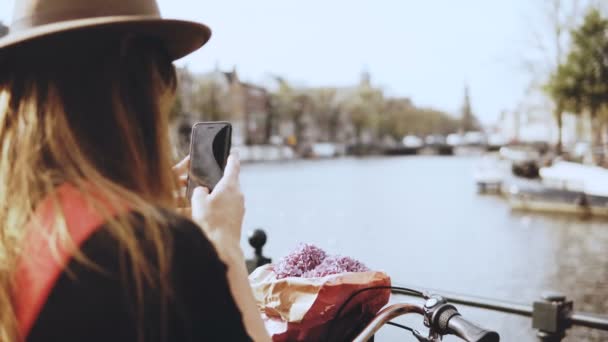 Dame touristique avec vélo prend des photos sur le pont. Femme aux cheveux longs et fleurs photographies beau paysage de rivière . — Video