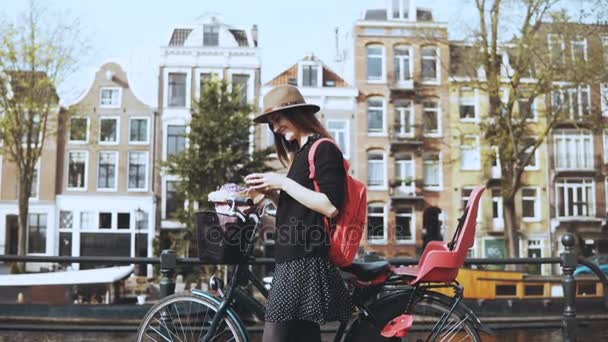 Söt flicka med cykel typer meddelande på bron. Kreativa fashionabla art arbetare på en bro med pittoreska gamla i stan. 4k — Stockvideo