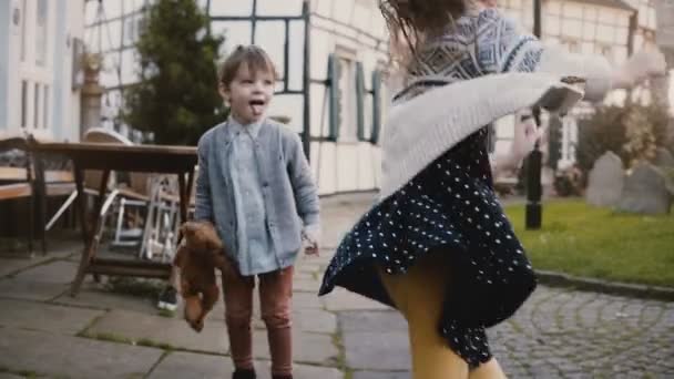 Zwei kleine Kinder machen Gesichter und tanzen draußen. Kaukasische Mädchen und Jungen mit Teddybär. Fachwerkhäuser Hintergrund 4k. — Stockvideo