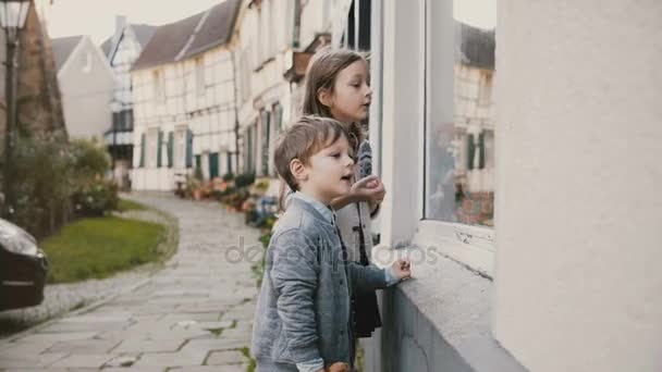 Κορίτσι και αγόρι, δείχνοντας δώρα σε παράθυρο κατάστημα παιχνιδιών. Δύο ευρωπαϊκές τα παιδιά κάνουν τις ευχές τους στο κατάστημα. Μισή ξυλεία τα σπίτια. 4k. — Αρχείο Βίντεο