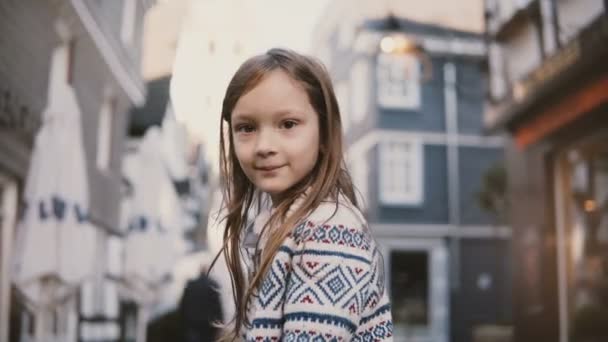 5-7 éves európai kislány portréja. Pózol a kamera mosolyogva, csodálatos, hosszú haj. Régi házak háttér. 4k. — Stock videók