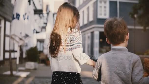 Camera volgt klein meisje en jongen lopen samen. Achteraanzicht. Twee kinderen zwerven rond de oude stad. Broer en zus. 4k. — Stockvideo