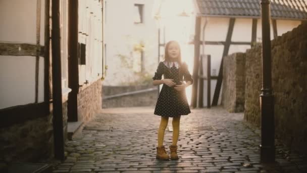 Πορτραίτο του χαριτωμένο κορίτσι Καυκάσιος πνέει τα φιλιά. Ευτυχισμένη γυναίκα παιδί θέτοντας σε φωτογραφική μηχανή σε ένα πλακόστρωτο δρόμο. Ηλιακό φως αναλαμπής. 4k. — Αρχείο Βίντεο