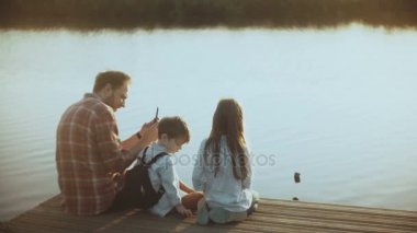 Baba ve iki çocuk birlikte bir göl iskelede oturup. Çocuk fotoğrafları Sukuşları smartphone ile Avrupa adamla. 4k.