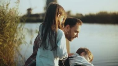 Sessiz gün batımı göl yakınındaki mutlu Beyaz aile sit. İki çocukla tek baba birlikte mutlu zaman harcamak. 4 k yakın çekim.
