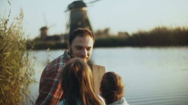 Hombre, niño y niña se sientan cerca de un lago al atardecer. Padre con dos hijos disfrutan del tiempo juntos. Relación familiar. 4K . — Vídeo de stock