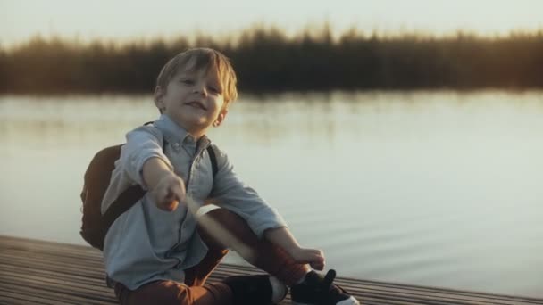Bedårande pojke sitter med en pinne på sjön pier. Porträtt av lilla kaukasiska barn pratar, tittar på kameran med glädje. 4k. — Stockvideo