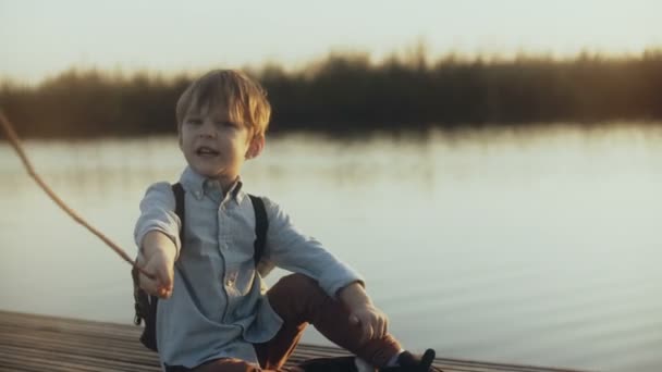 Милый мальчик сидит с палкой возле закатного озера. Портрет кавказского ребенка, играющего злого дикого пирата на пирсе. 4K . — стоковое видео