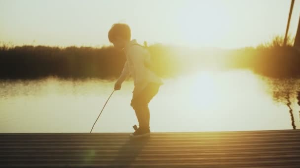 Парень играет с палкой на пирсе у озера на закате. Вид сзади. Маленький исследователь. Удивительный горизонт вспышки солнечного света. 4K . — стоковое видео