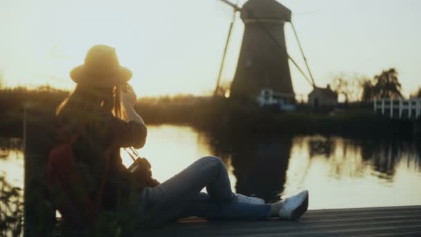 Mujer con sombrero se sienta en el muelle del lago al atardecer Holanda. La blogger con smartphone y cámara toma una foto del molino de viento. 4K . — Vídeos de Stock