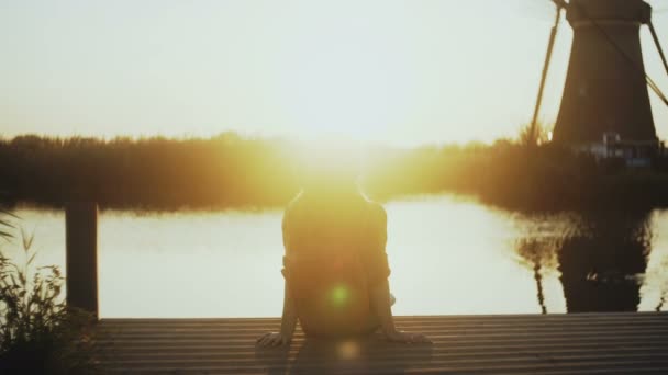 Una donna si siede su un piccolo molo del lago, guardando il tramonto. Ora d'oro. Momento perfetto di pace e armonia interiore. Vista posteriore 4K . — Video Stock