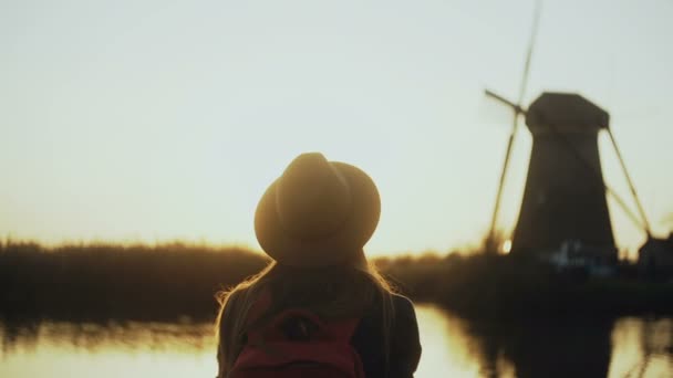 Gärna kvinnliga resenären i hatt klockor nedgående solen. Kvinnan ler på en vacker sjö brygga, gamla wind mill. 4k tillbaka Visa. — Stockvideo