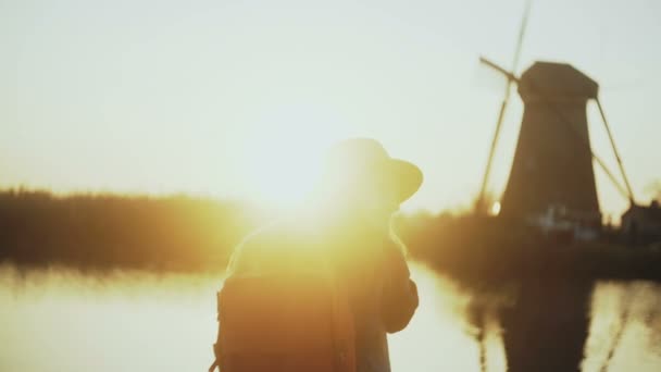 Γυναίκα με καπέλο κάθεται στην προβλήτα ήσυχο ηλιοβασίλεμα στη λίμνη. Κοιτάζει δεξιά, παίρνει το τηλέφωνό της. Παραδοσιακό ολλανδικό ανεμόμυλο. Δείτε 4k πίσω. — Αρχείο Βίντεο