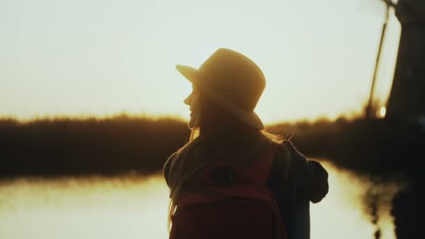 Felice ragazza carina in cappello si siede sul molo del fiume tramonto. Ora d'oro. Godendo meraviglioso momento di viaggio. 4K vista posteriore primo piano . — Video Stock