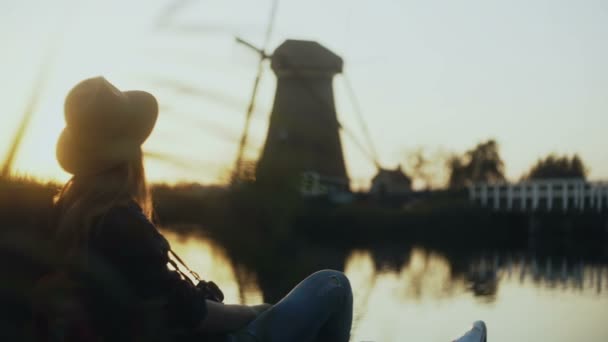 Nöjd resenär kvinna i hatt solnedgången. Flickan sitter på en vacker sjö brygga med kamera. Rustika wind mill. 4k. — Stockvideo