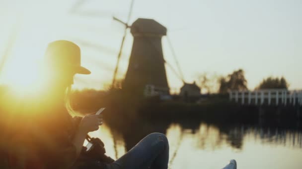 Flicka i hatt sitter på sunset landsbygdens sjön pier. Turist kvinna tar ett foto av gamla holländska väderkvarn med smartphone. 4k. — Stockvideo