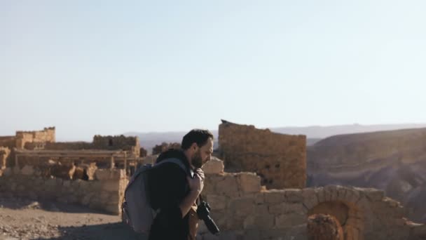 Professionell fotograf utforskar antika ruiner. Europeiska mannen med kameran och ryggsäck nära bergsutsikt. Israel 4k. — Stockvideo