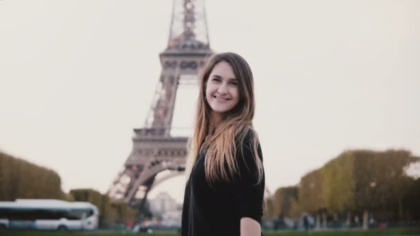 Gelukkig jongedame permanent in de buurt van de Eiffeltoren in Parijs, Frankrijk, glimlachend op de camera en de klap kus verzenden. — Stockvideo