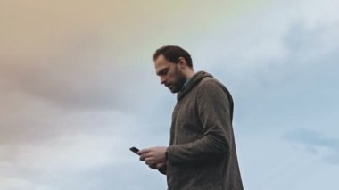 Sakallı Avrupa adam smartphone ile duruyor. Düşük açılı şaşırtıcı gökyüzü görünümü. Casual yakışıklı erkek smartphone fotoğrafı çeker. 4k