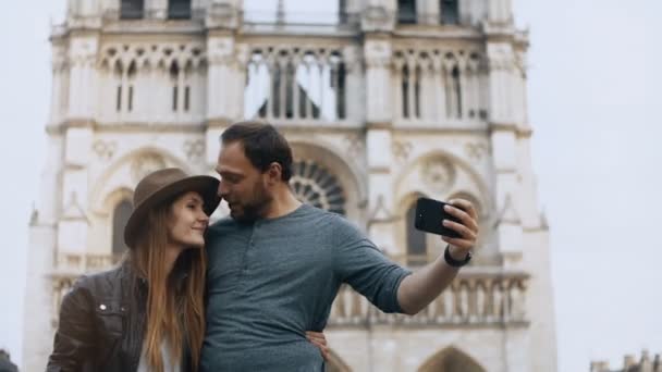 Joven pareja feliz abrazando y tomando fotos selfie en el teléfono inteligente cerca de la famosa Notre Dame en París, Francia . — Vídeos de Stock