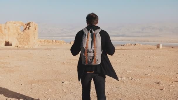 Man with backpack walks on desert mountain top. Relaxed European male tourist enjoys Dead Sea panorama. Israel 4K. — Stock Video