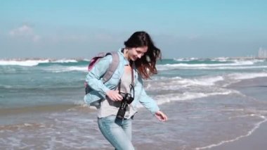 Güzel beyaz kız sahilde dalgalar çalışır. Muhteşem bir kadın kamera gülümsüyor. Saç esen rüzgar. Ağır çekim.