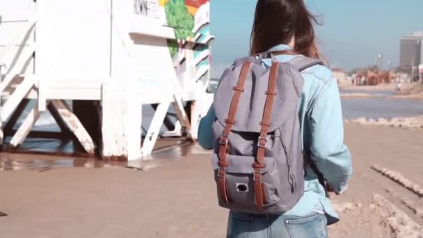 Ragazza con zaino grigio cammina su una spiaggia. Primo piano. Capelli che soffiano nel vento. Posto di guardia costiera e case. Rallentatore . — Video Stock