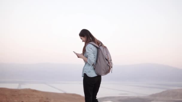 Vrouw met behulp van smartphone in de buurt van schemering montain landschap. Slow-motion. Casual meisje met rugzak typen in messenger-app. Israël. — Stockvideo