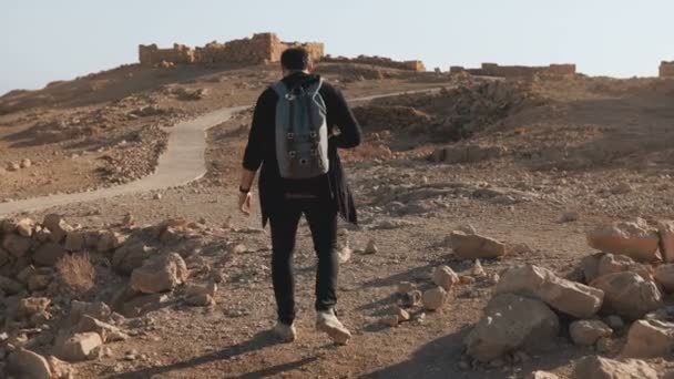 Man with backpack hiking among ancient ruins. Relaxed European male traveler walks on desert rocks and sand. Israel 4K. — Stock Video