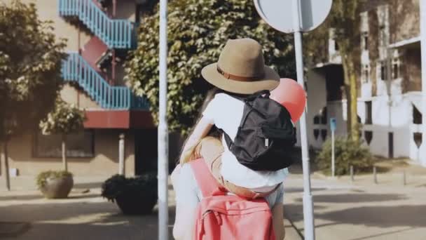 Madre lleva a su hijo en hombros entre las casas. Mujer caminando con un niño en sombrero y dos globos aéreos. Estilo de vida 4K . — Vídeos de Stock