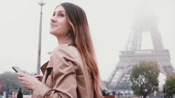 Joven mujer hermosa usando el teléfono inteligente cerca de la torre Eiffel. Mujer sonriente mirando a su alrededor, mira París, Francia . — Vídeos de Stock