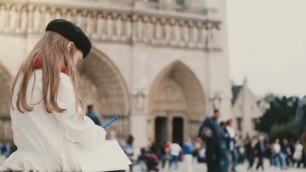 Linda niña en boina sentada en un lugar lleno de gente y dibujo. Niño morena cerca de la de Notre Dame en París, Francia . — Vídeos de Stock