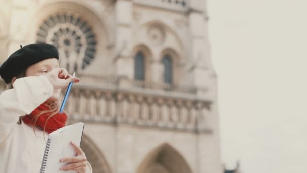 Mała dziewczynka stylowy Beret rysunek obrazu w notesie w pobliżu katedry Notre Dame w Paryżu, robi jej hobby. — Wideo stockowe
