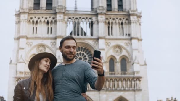 Joven pareja feliz tomando fotos selfie en el teléfono inteligente cerca de la Notre Dame en París, Francia, disfrutando de la hermosa vista . — Vídeos de Stock