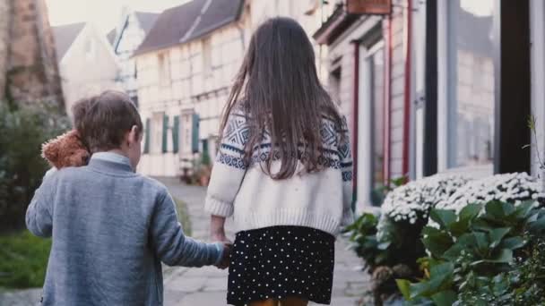 La cámara sigue a dos niños pequeños caminando tomados de la mano. Vista trasera en cámara lenta. Niña y niño vagan por el casco antiguo. ¡Hermanos! . — Vídeos de Stock