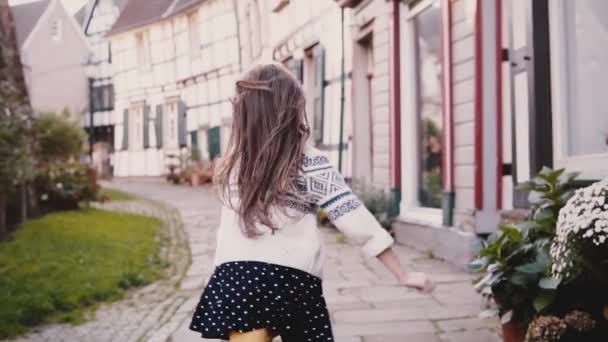 Meisje kijkt naar camera, draait op verharde weg. Achteraanzicht. Slow-motion. Schattig Europese kind uitgevoerd in oude straat. — Stockvideo