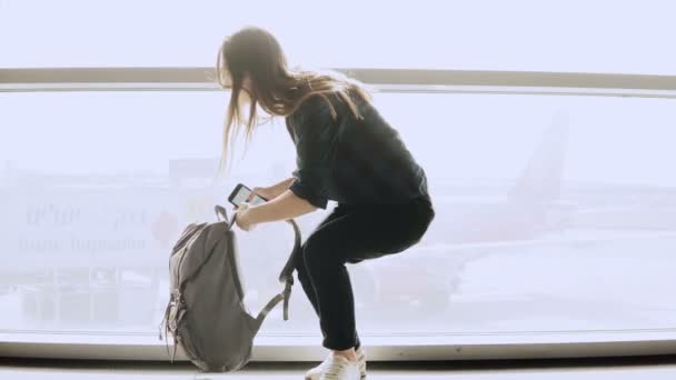 Mujer feliz se sienta con teléfono inteligente por ventana del aeropuerto. Chica caucásica con mochila utilizando la aplicación de mensajero en la terminal. 4K . — Vídeo de stock