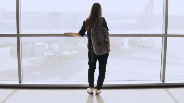 Mujer joven con mochila cerca de la ventana terminal. Turista femenina caucásica usando smartphone en el salón del aeropuerto. Viajar. 4K . — Vídeo de stock
