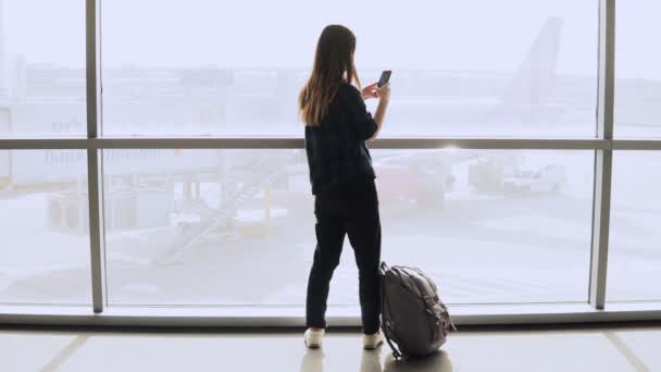 Chica toma selfie cerca de la ventana del aeropuerto, se aleja. Feliz turista caucásica usando smartphone en terminal hall. 4K . — Vídeo de stock