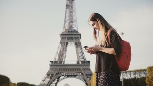 Young beautiful woman with backpack standing near the Eiffel tower and using the smartphone, browsing the Internet. — Stock Video