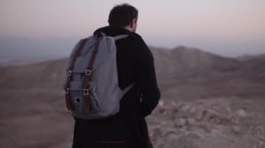 Adam sırt çantası ile alacakaranlık Çölü'hitchhikes. Ağır çekim. Smartphone autostops yakınındaki bir gün batımı karayolu ile beyaz erkek