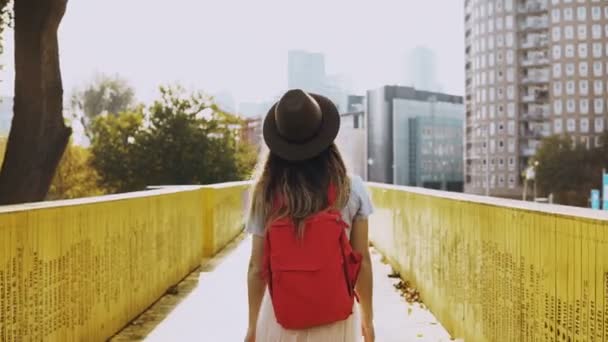 Menina de chapéu com longos passeios de cabelo em uma ponte de cidade. Mulher branca entre paredes amarelas com mochila vermelha. Visão traseira 4K . — Vídeo de Stock