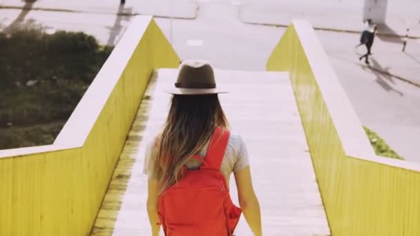 Una donna con i capelli lunghi cammina su un ponte cittadino. Ragazza europea con zaino rosso. Pareti gialle della passerella urbana. 4K . — Video Stock