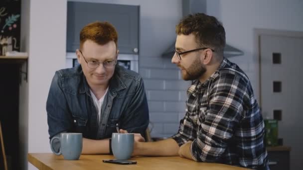Dos amigos varones se sientan junto a la mesa de la cocina y charlan. Los hombres caucásicos comparten una conversación amistosa con el café en casa. 4K . — Vídeo de stock
