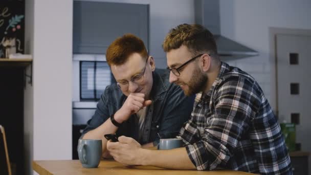 Dos amigos varones ven videos divertidos en línea. Guapos hombres europeos felices usando teléfono inteligente con café en la cocina. 4K . — Vídeos de Stock