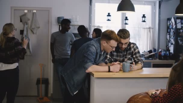 Two guys use smartphone app at office kitchen. Happy young European men using social networks at a diverse party. 4K. — Stock Video