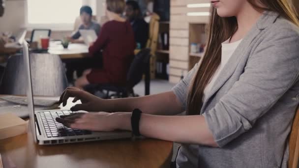 Une jeune fille européenne concentrée tapant une lettre sur un ordinateur portable. Travail acharné dans un espace de bureau branché, travail d'équipe en arrière-plan 4K . — Video