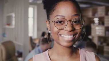Oldukça genç Afro-Amerikan mutlu kadın lider kamera modern ofis arka 4 k ortak çalışma içinde gülümseyen bardakları