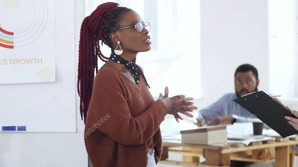 Happy young casual African woman boss in eyeglasses leading discussion, chatting to colleagues at modern light office.