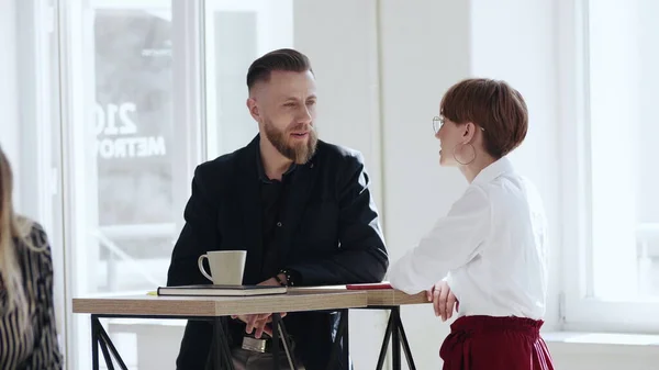 Feliz sonriente barbudo de mediana edad jefe masculino hablando con la joven colega mujer de pie en la mesa de oficina loft de moda . — Foto de Stock